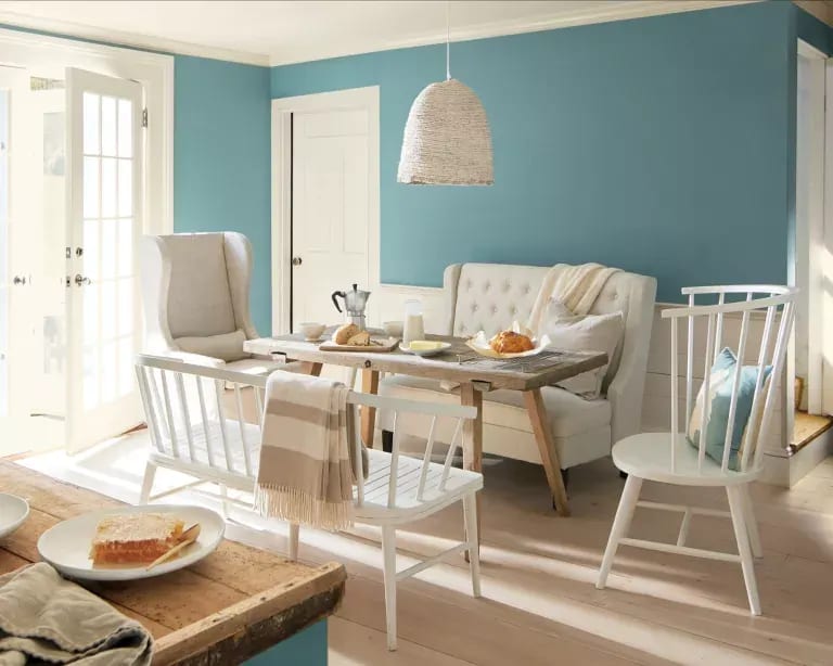 Dining nook with a white banquette, rustic wood table, woven pendant, and teal accent wall.