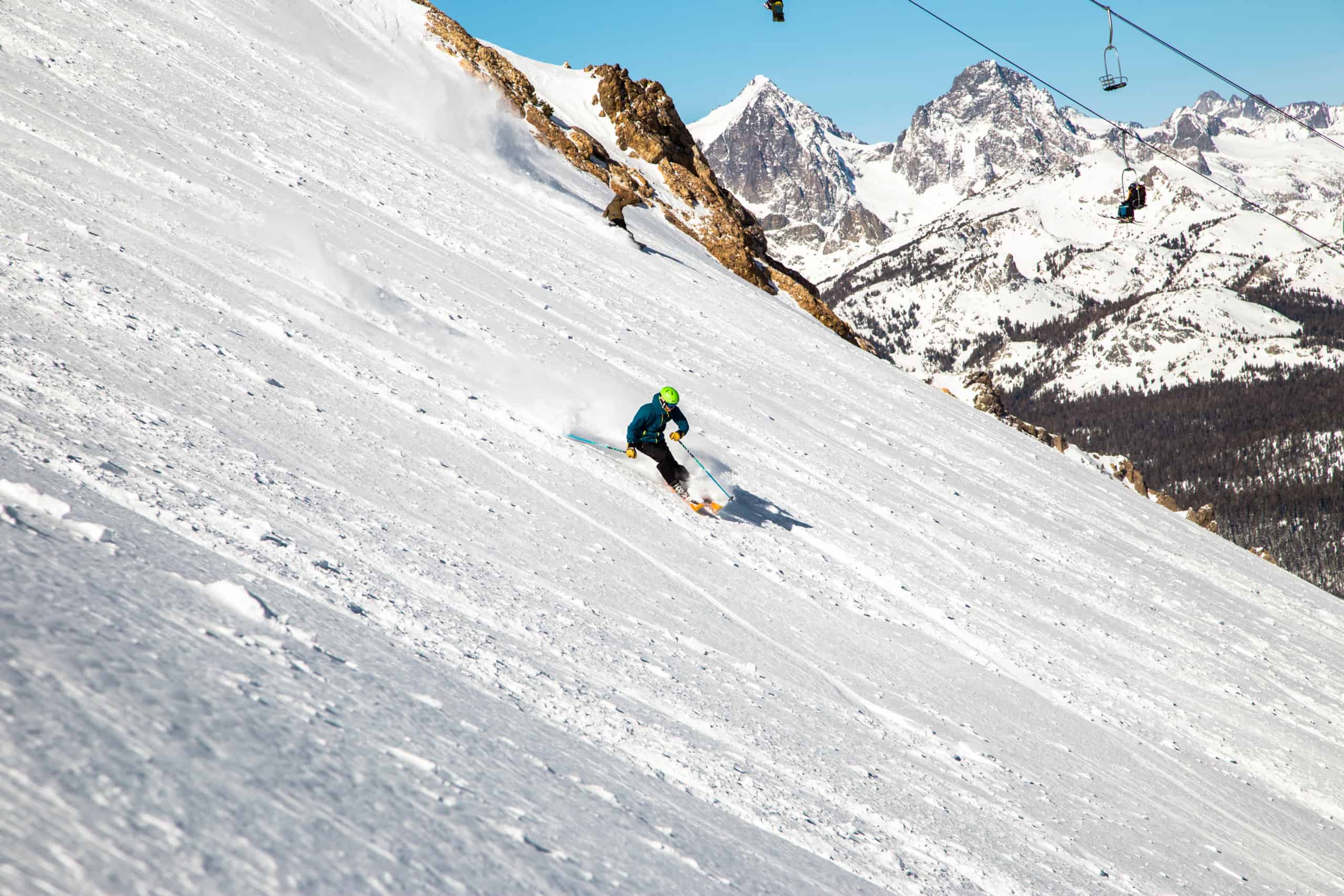 Skier in a green helmet makes a turn down a steep hill on Mammoth Mountain with Banner Peak and Ritter Peak and a chairlift in the background
