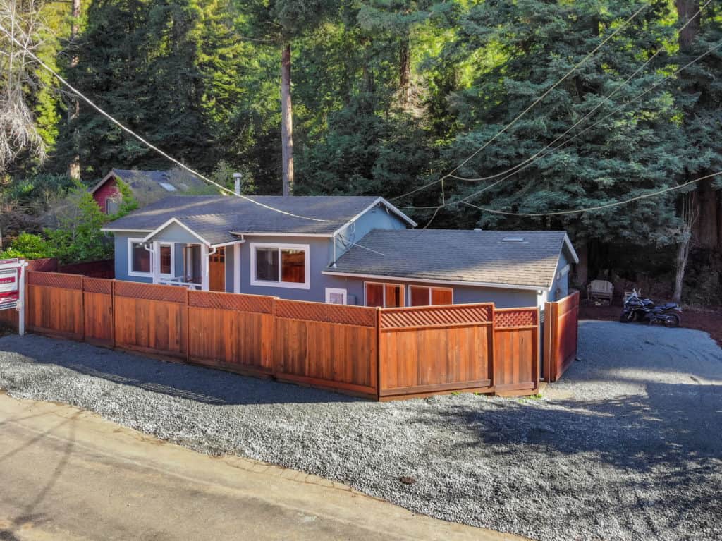 Blue single-story home behind a reddish wood fence, with dense woods behind it.