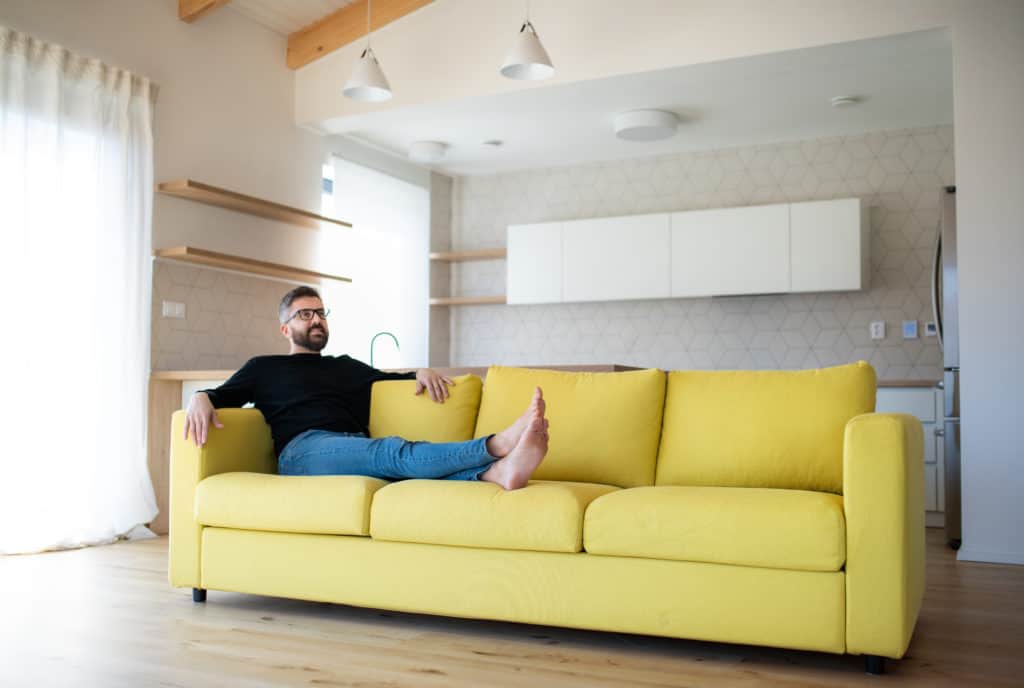 Man relaxing barefoot on a bright yellow sofa in a light-filled modern kitchen.