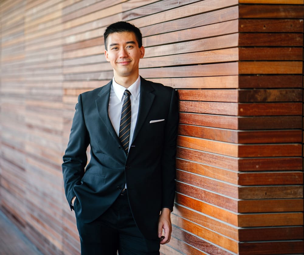 Man in a dark suit and striped tie leaning against a wooden slat wall.