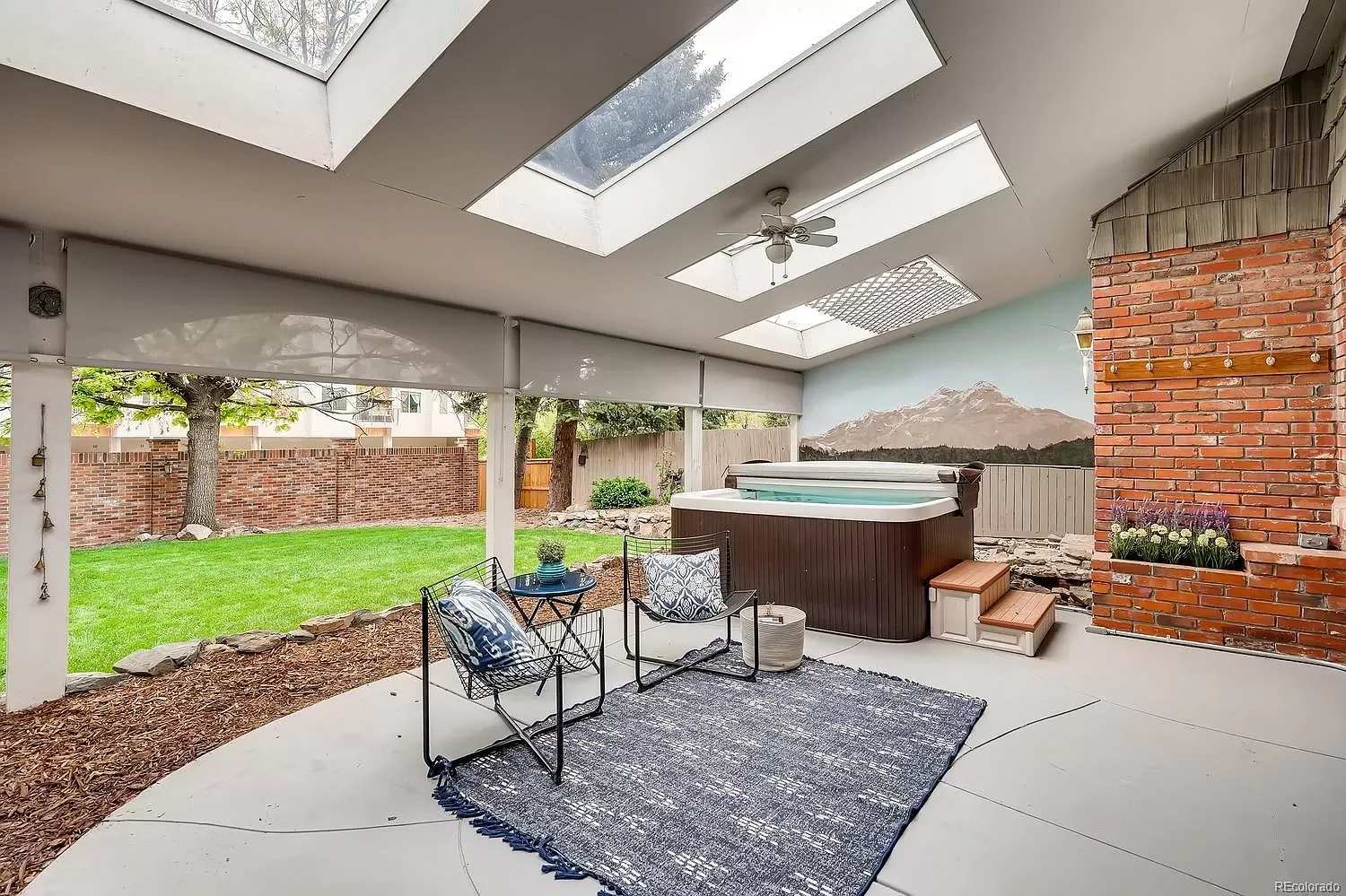 Covered patio with skylights, a hot tub, and brick fireplace beside a grassy backyard.