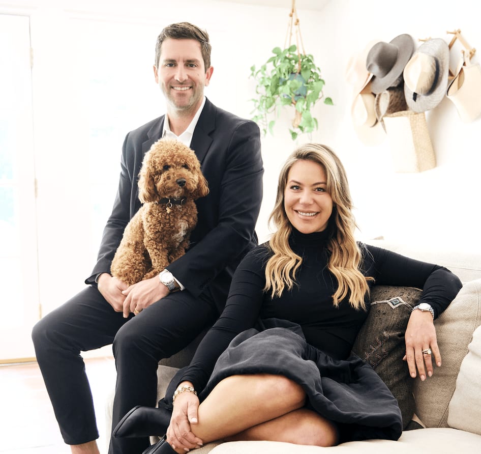 Spencer Sanderson and Heather Sanderson smile happily with a brown dog in the living room.