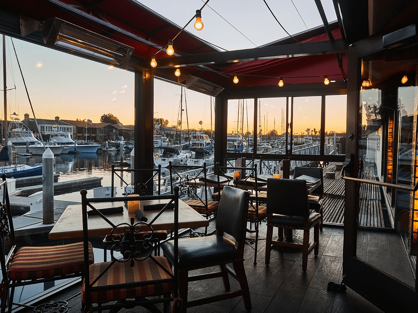 Waterfront restaurant dining area with sunset view and docked boats visible through large windows.