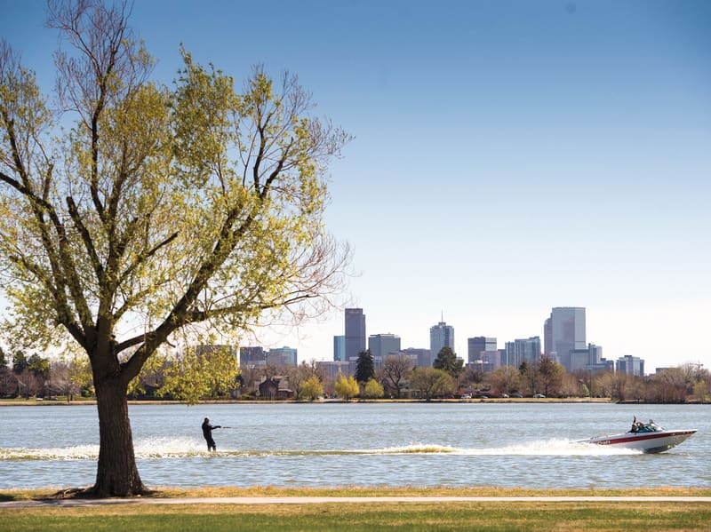 Denver skyline across a lake, with a water-skier and motorboat beneath a large autumn tree.