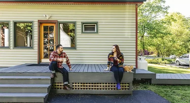 Two people sitting on the porch of a small house, enjoying each other’s company. The setting is casual and relaxed, indicating a comfortable home environment.