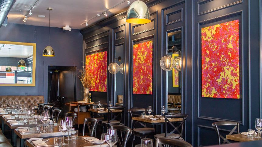 Photo of the interior of Seven Hills Restaurant in Russian Hill, San Francisco. The restaurant is dimly lit and has a warm and inviting atmosphere. There are tables and chairs arranged throughout the space, and there is a bar in the back. The walls are adorned with artwork, and there are candles on the tables.