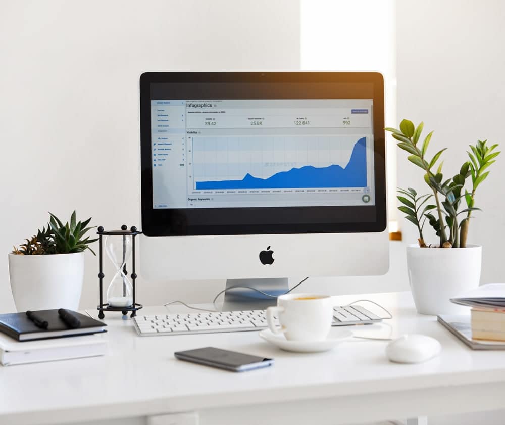 Home-office desk with a computer displaying a blue analytics graph, coffee, notebooks, and potted plants.