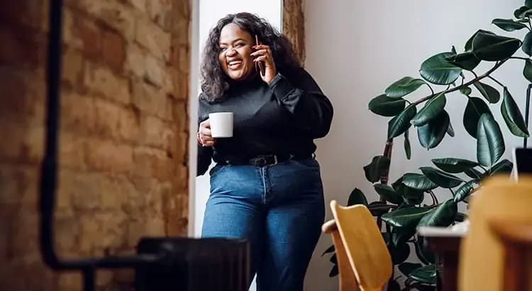 A woman holding a cup while talking on the phone and standing next to a plant. She is dressed casually and smiling, indicating a relaxed moment.