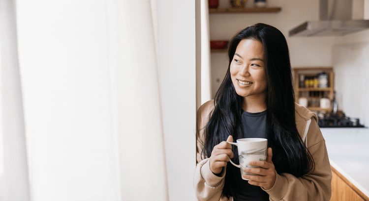 Woman holding a mug in a bright kitchen, looking toward the window.