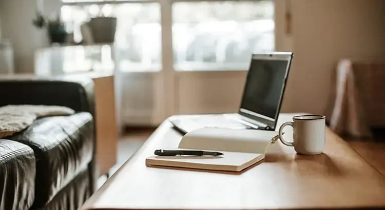 A home office setup with a laptop, coffee mug, and other office supplies on a wooden desk. The background is softly lit, suggesting a productive work environment.