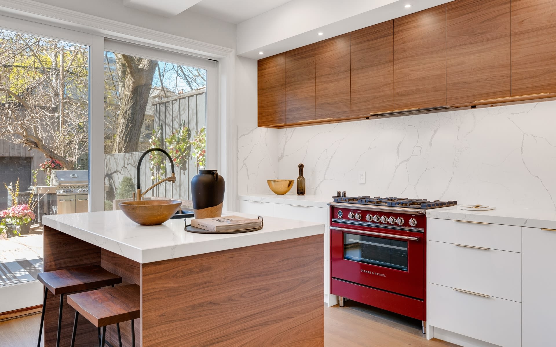Modern kitchen with a walnut island, marble counters, red range, and garden views.