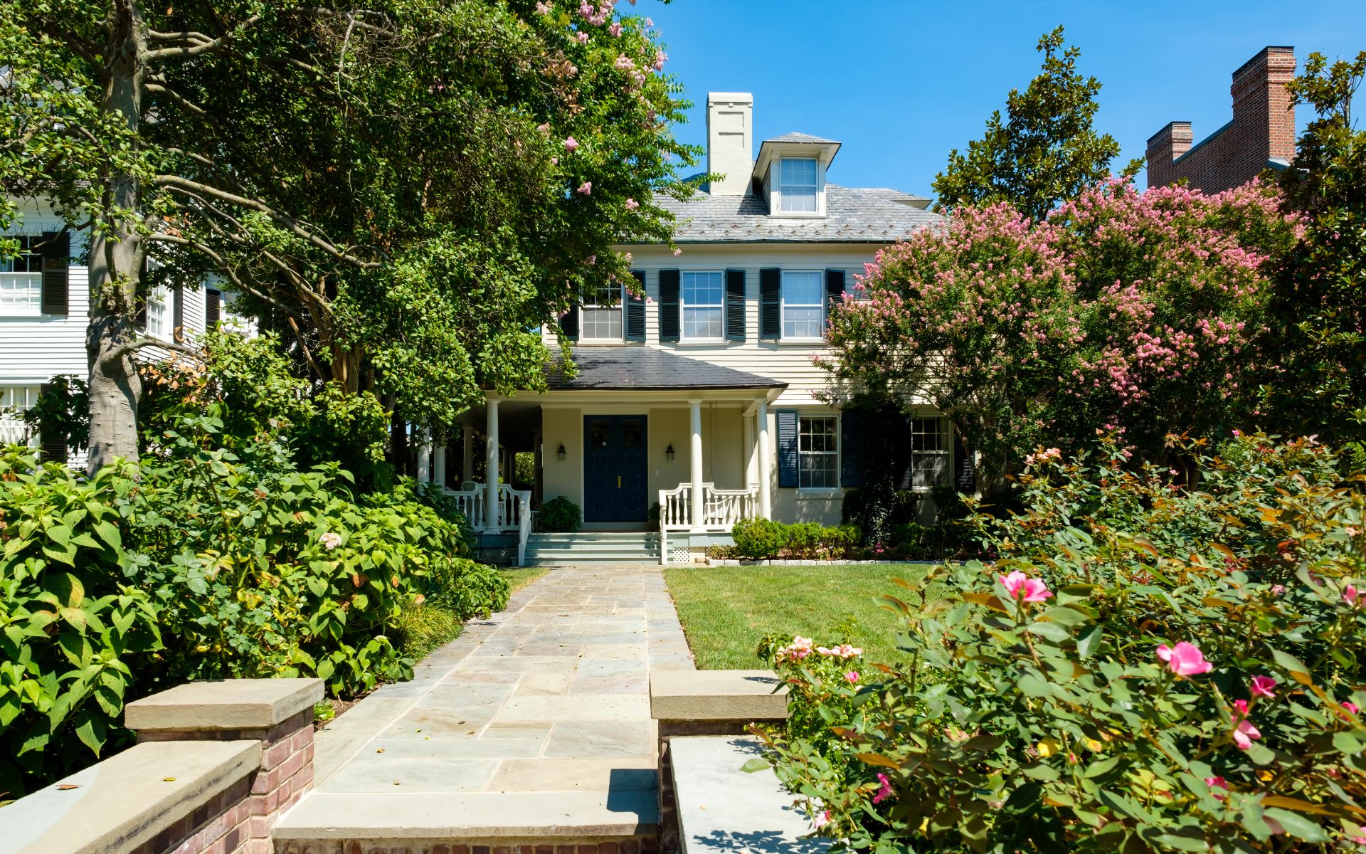 Two-story house with a covered porch, stone walkway, and flowering pink shrubs.