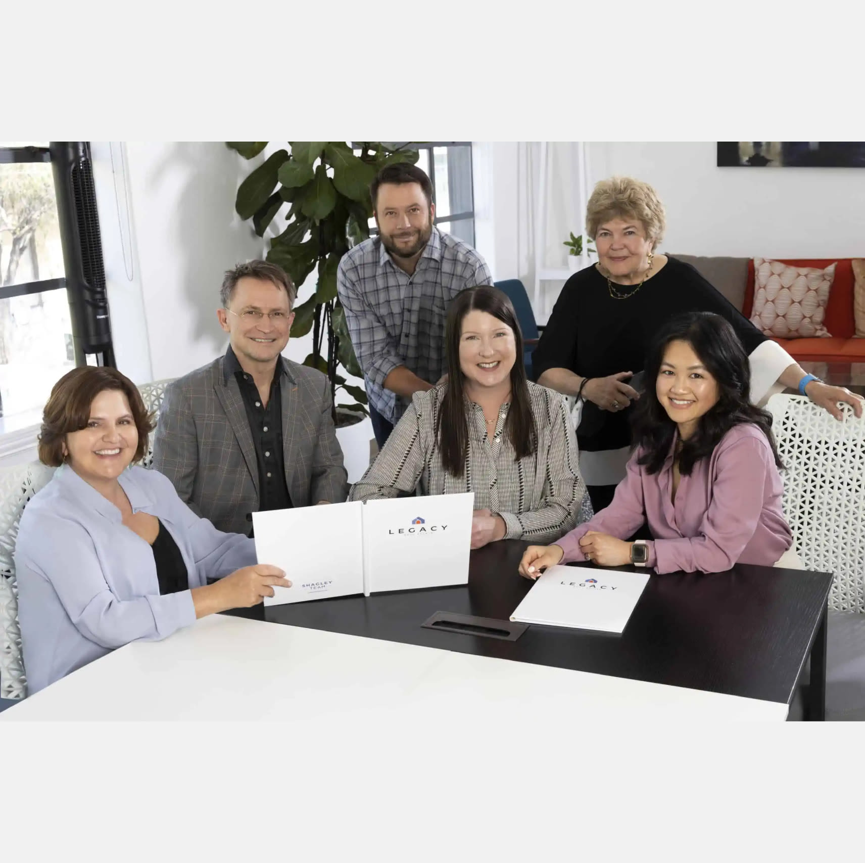 A professional and cohesive real estate team photo captured at a desk setting. The team members are seated together, showcasing unity and collaboration.  The image portrays a dedicated and knowledgeable real estate team, fully equipped to guide clients through the buying or selling process with expertise and care.