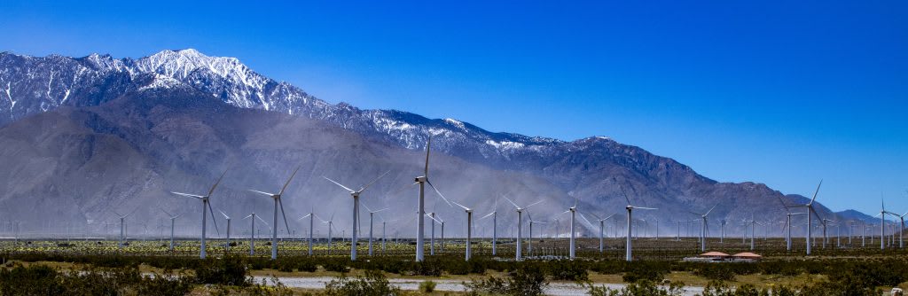 A panoramic view of wind turbines in a desert landscape, with snow-capped mountains in the background under a clear blue sky.
