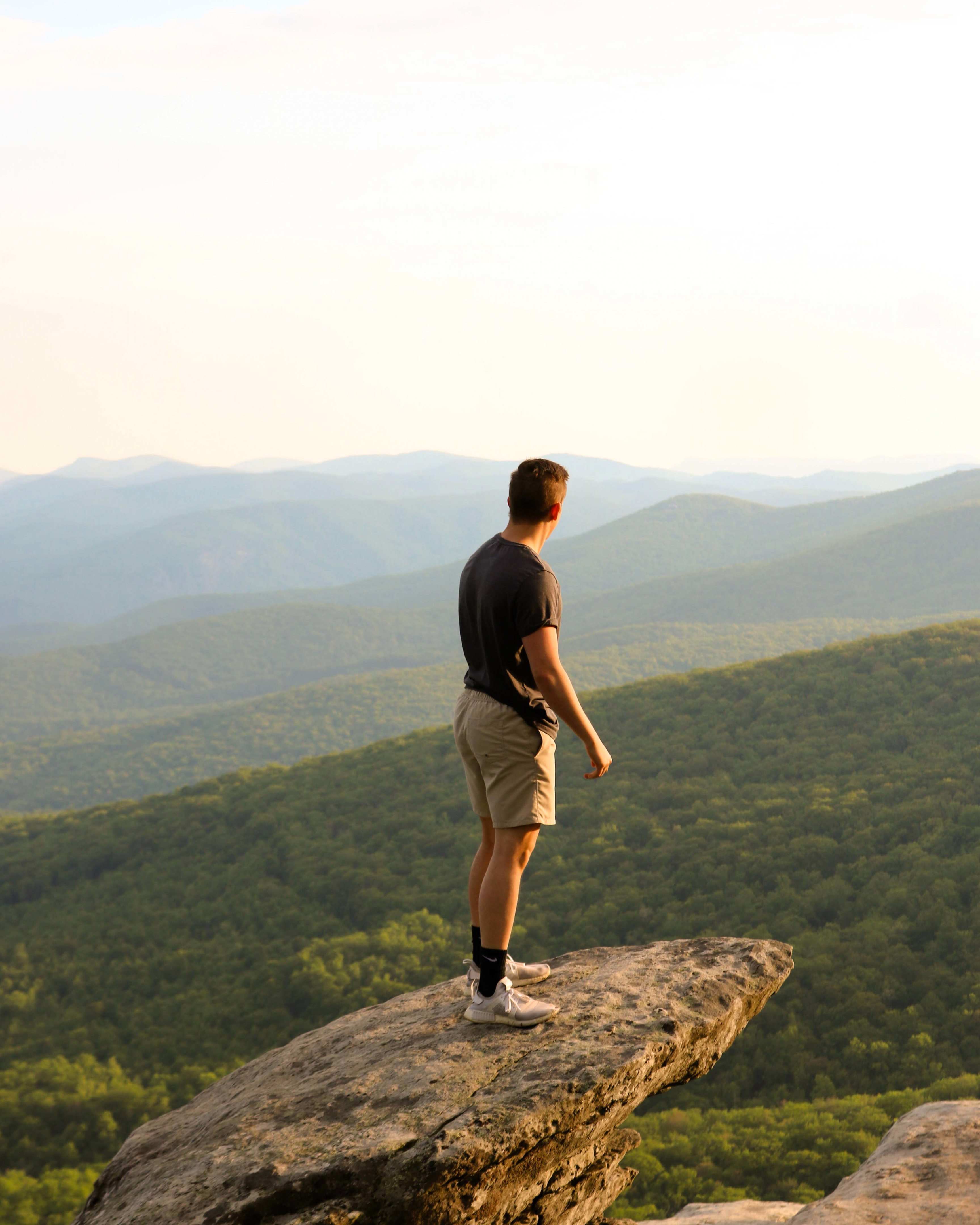 A person stands on a rocky cliff edge, an ideal platform for reflection, overlooking a vast landscape of green rolling hills under a clear sky.