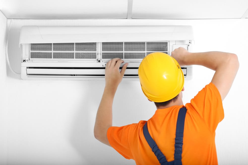 A worker in an orange shirt and yellow hard hat is servicing an air conditioning unit on a white wall.