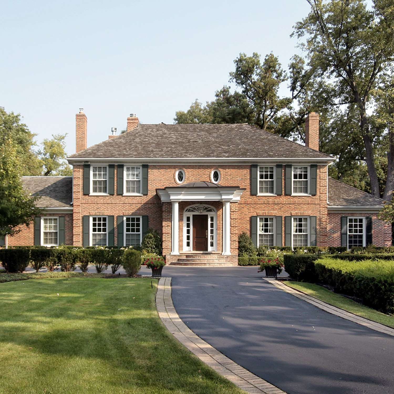 Red-brick house with a white-columned entrance portico, green shutters, and a curved driveway.