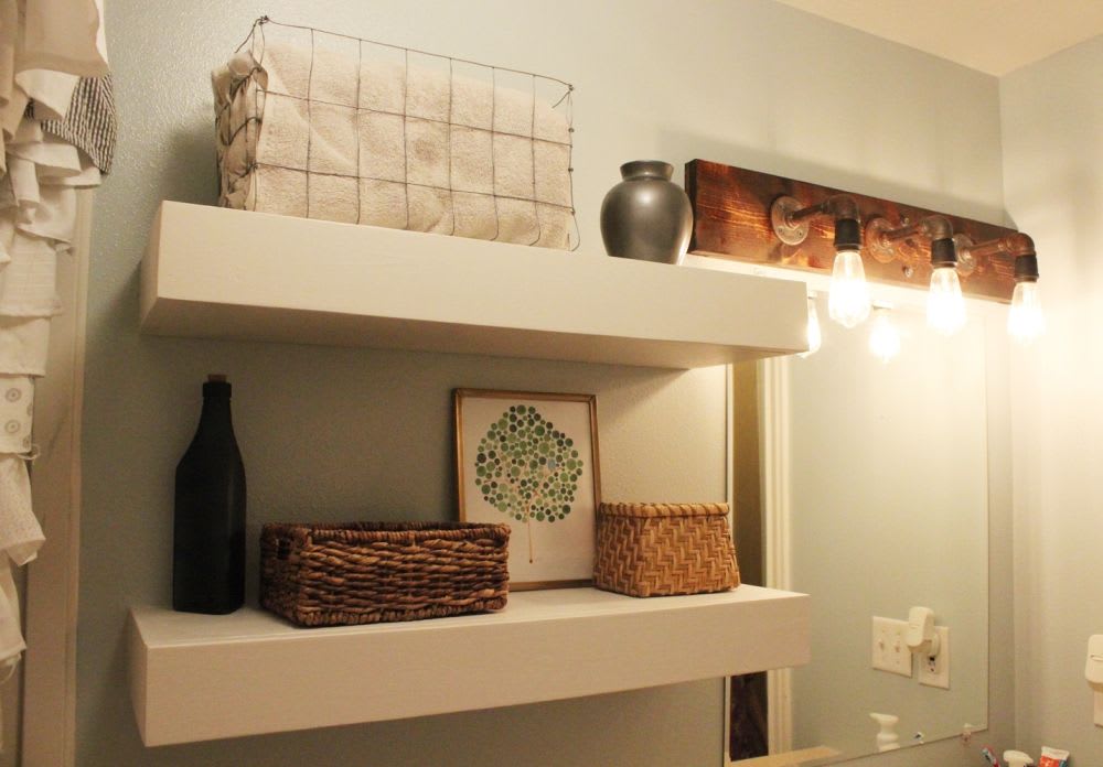 A bathroom shelf displays towels, decorative baskets, a vase, and a framed tree print, with a rustic light fixture above.