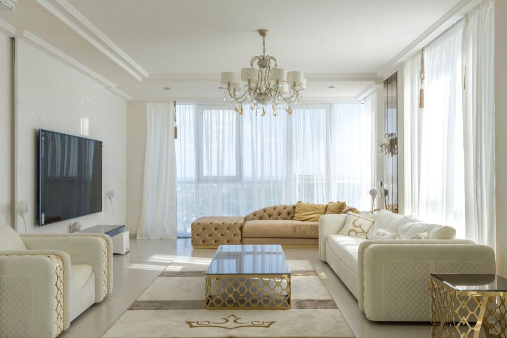 Luxurious living room with white sofas, tufted chaise lounge, chandelier, gold-accented furniture, and large windows.