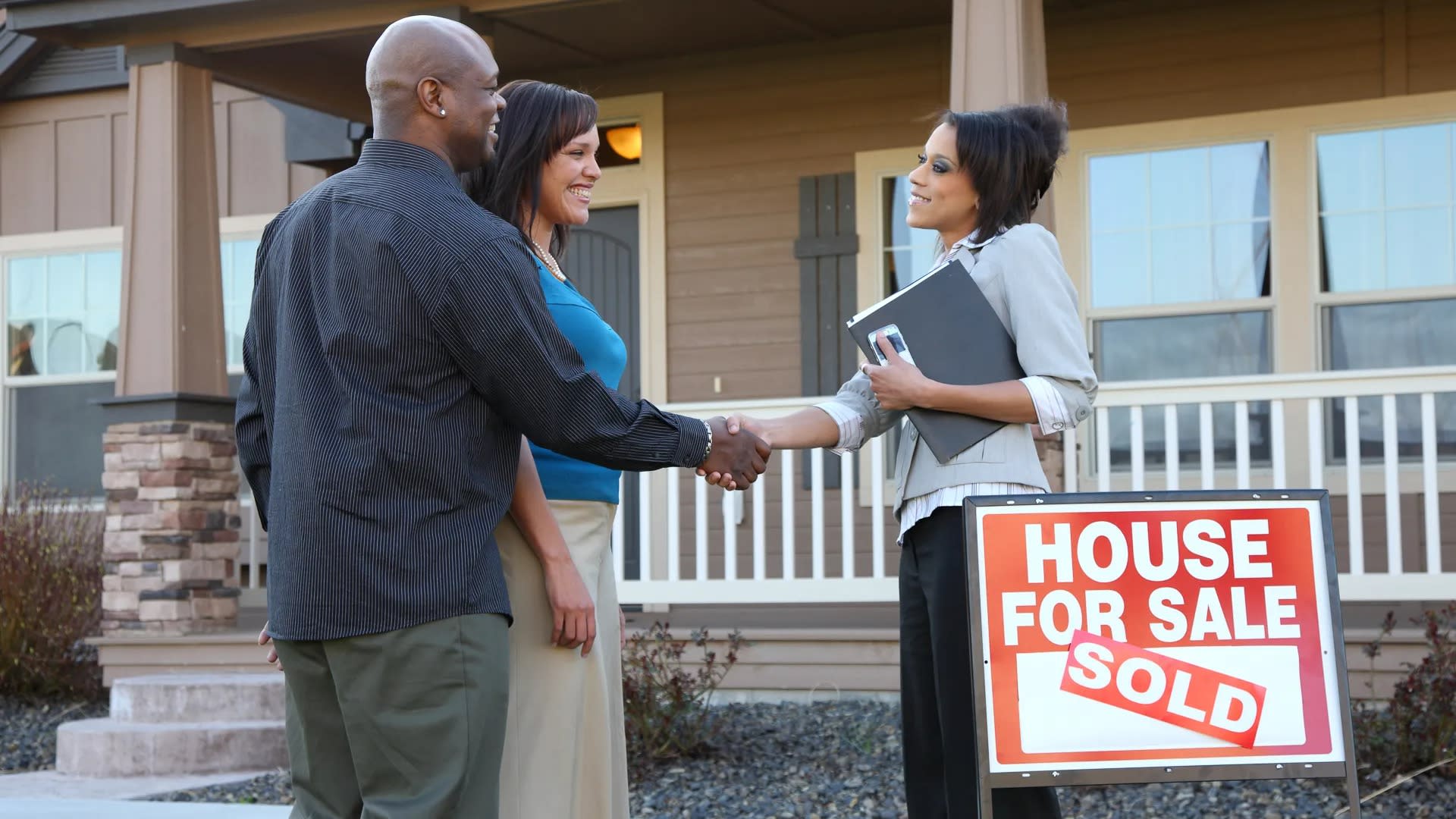 Real estate agent shaking hands with a couple in front of a sold house with a 'House For Sale Sold' sign.