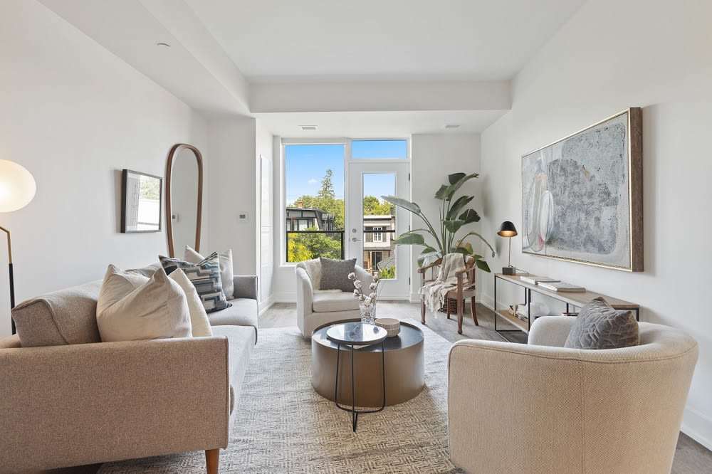 Bright living room with neutral seating, a round coffee table, and large windows overlooking trees and neighboring homes.