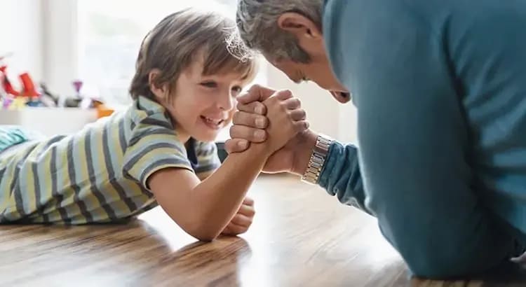 a child arm-wrestling with an adult, likely a playful interaction between a parent and child. They are smiling and enjoying the moment.