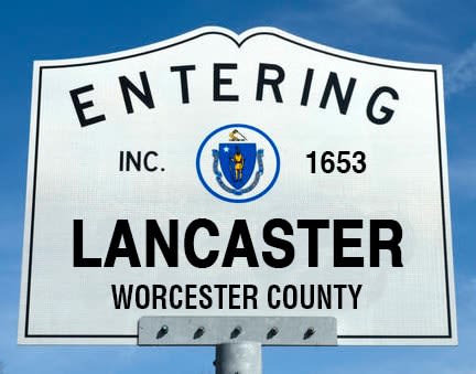 White street sign for Lancaster, Massachusetts, under a clear blue sky. 