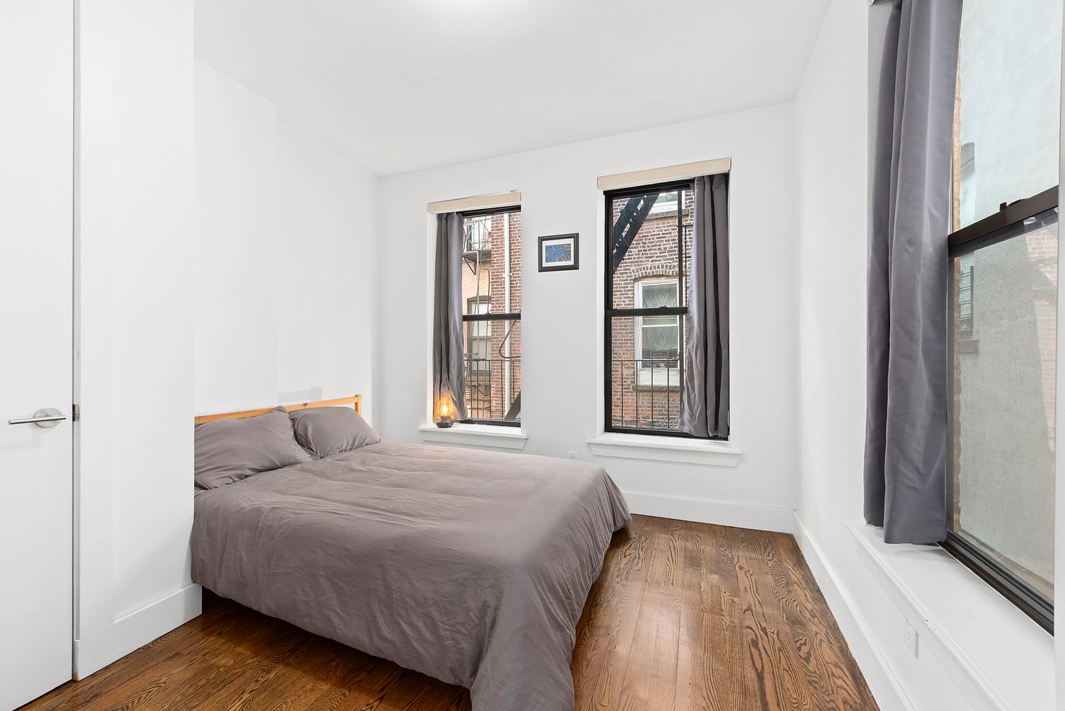 Bedroom with a gray-covered bed, hardwood floors, and tall windows facing neighboring brick buildings.