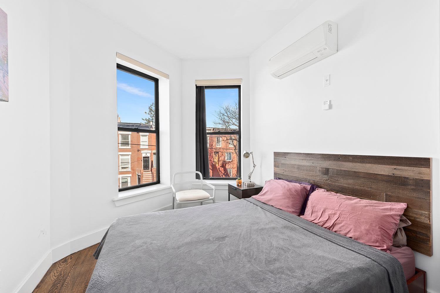 Bright bedroom with gray bedding, pink pillows, a wood headboard, and two tall windows.