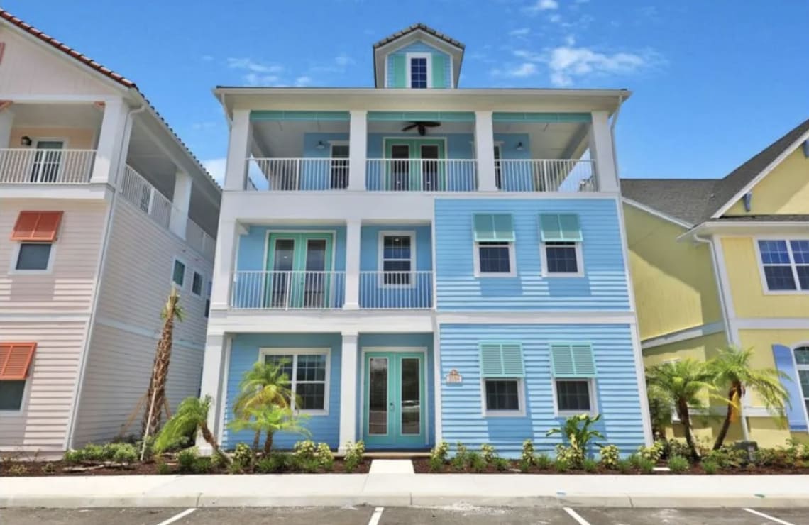 A blue and white two-story house with a balcony, a walkway, flower beds, trees, and other homes in the background.