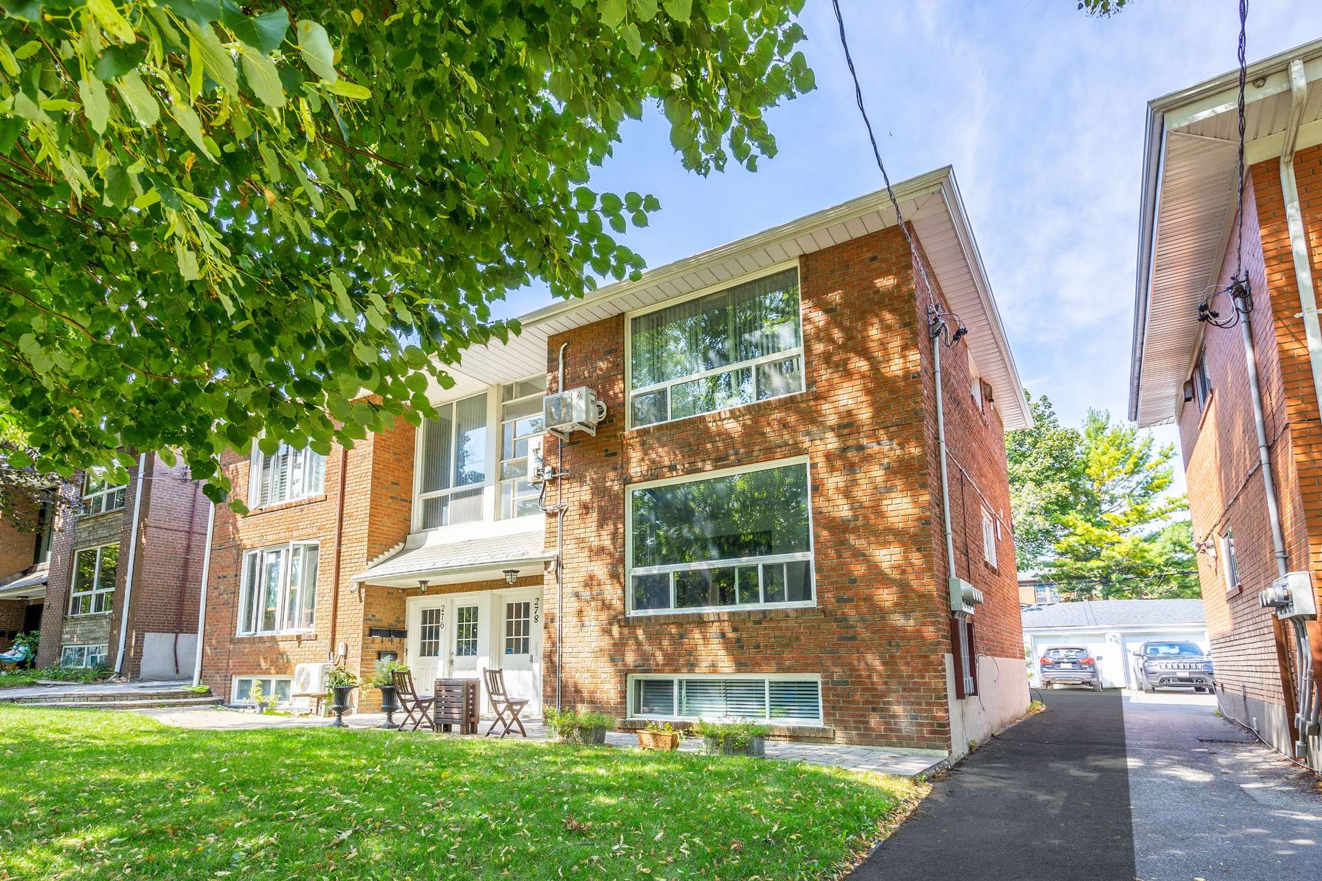 Red-brick multi-unit building with large windows, leafy shade, a small lawn, and driveway.