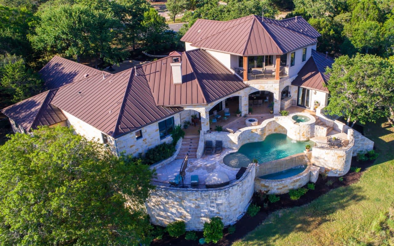 An aerial view of a large, red-brick house with a large, blue pool. The sky is a clear blue and the sun is shining.