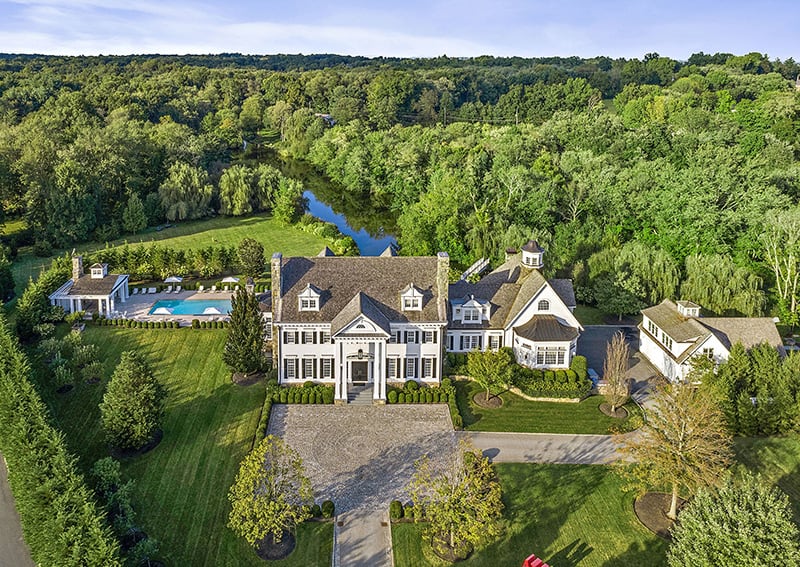 Aerial view of a white estate with a swimming pool, landscaped grounds, and surrounding woodland.