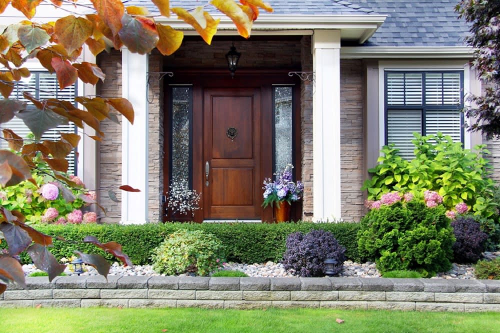 A house with a wooden door and a lush green lawn, white door posts, black roof, and well groomed plants.