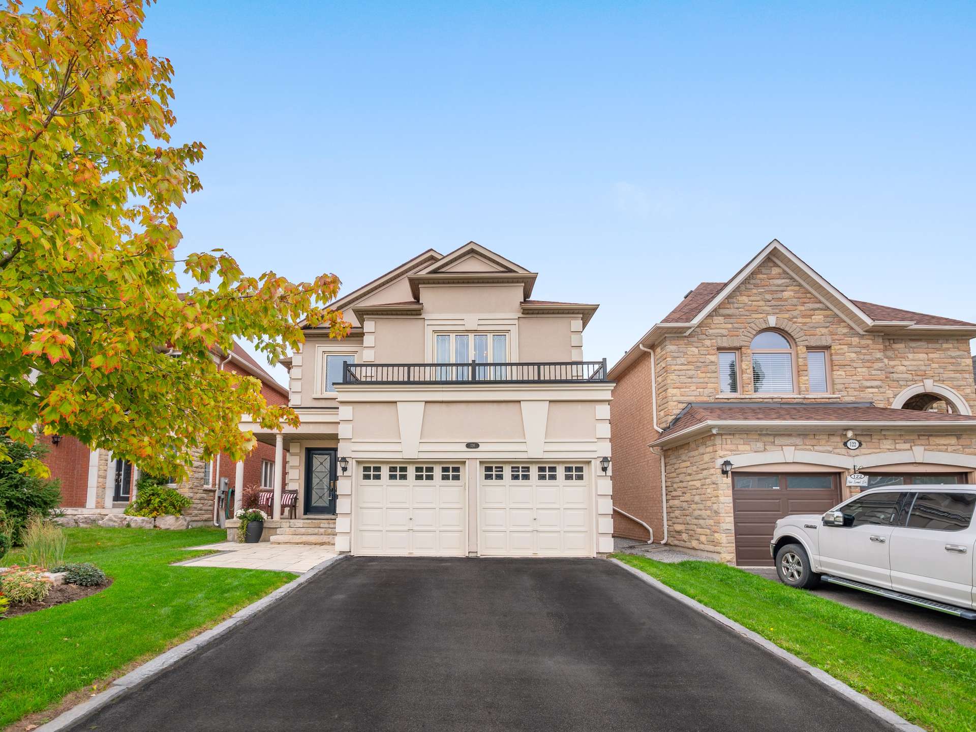 Two-story beige house with a double garage, balcony, and paved driveway beneath autumn leaves.