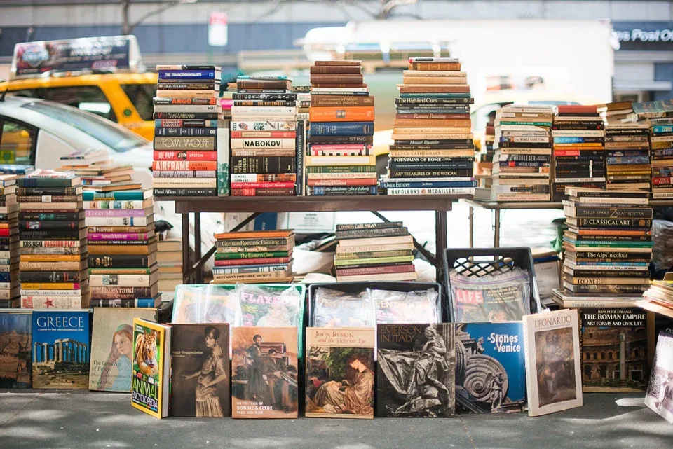 Stacks of used books at a sidewalk bookstall, with colorful covers displayed along the pavement.