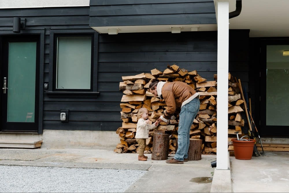 A man and a child interact outside a modern home, with a stack of firewood in the background.