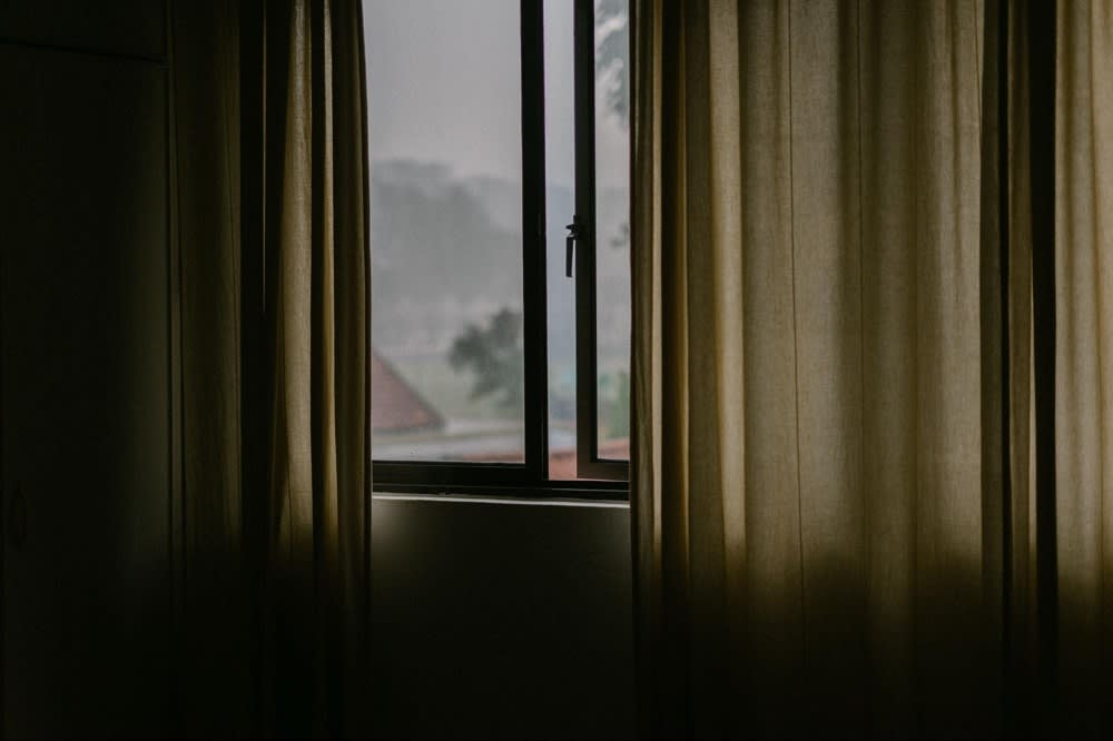 A dimly lit room with sheer curtains framing a window, revealing a cloudy, overcast view outside.