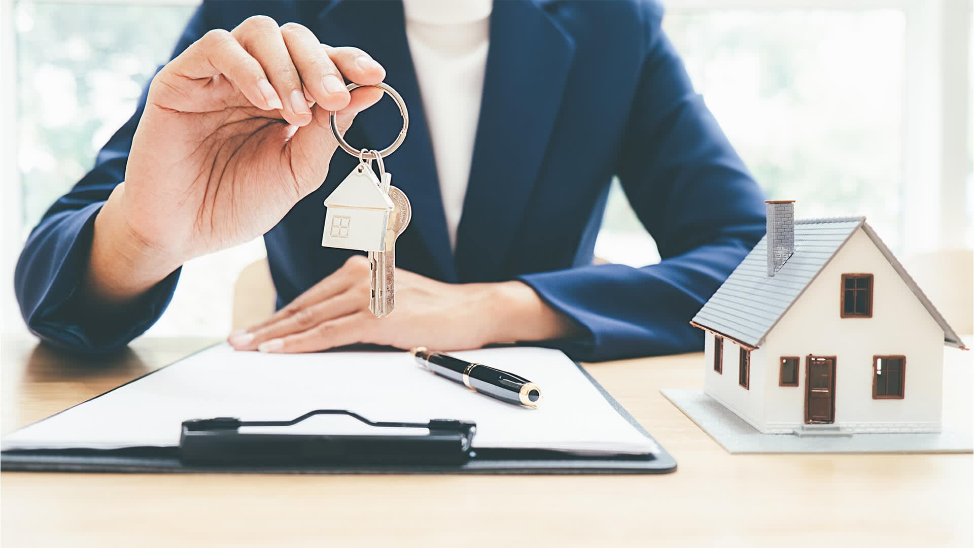 Real estate agent holding keys to a house over a contract with a model house on the desk.