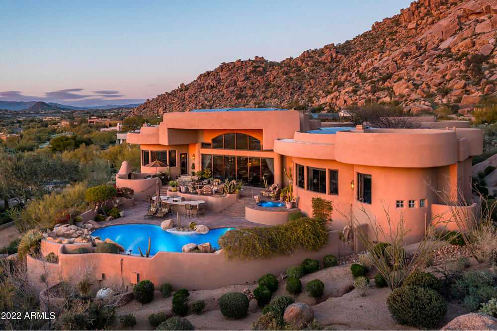 Luxury desert home with curved architecture, large windows, and a swimming pool nestled against a rocky hillside.