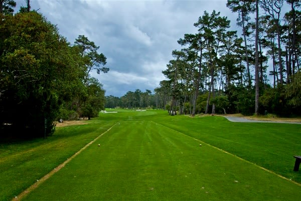 Golf course fairway from the tee, bordered by mature trees beneath a cloudy sky.