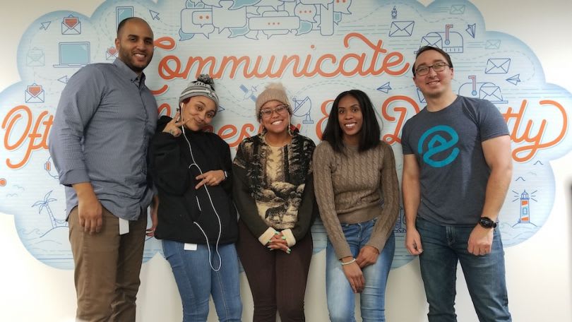 Five coworkers pose in front of a colorful communication-themed mural.