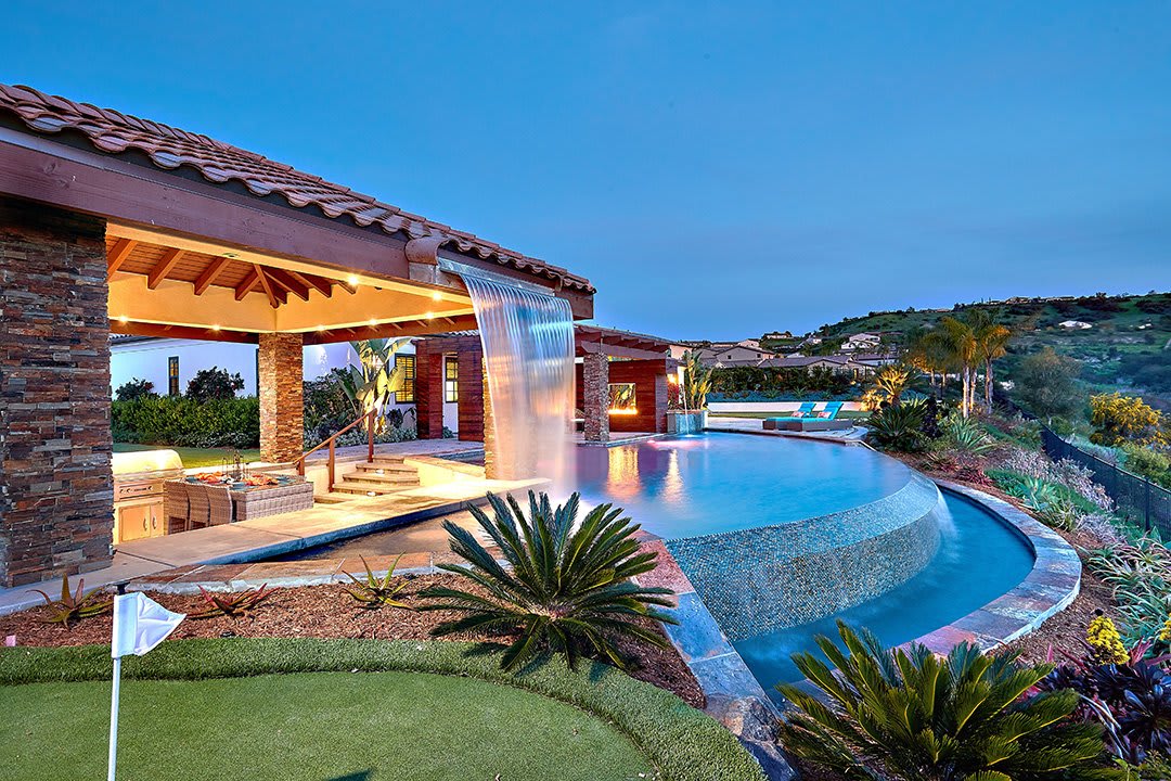 Side view of backyard with putting green, waterfall edge on infinity pool, covered patio with sunken entertaining area and rooftop waterfall and golf course views