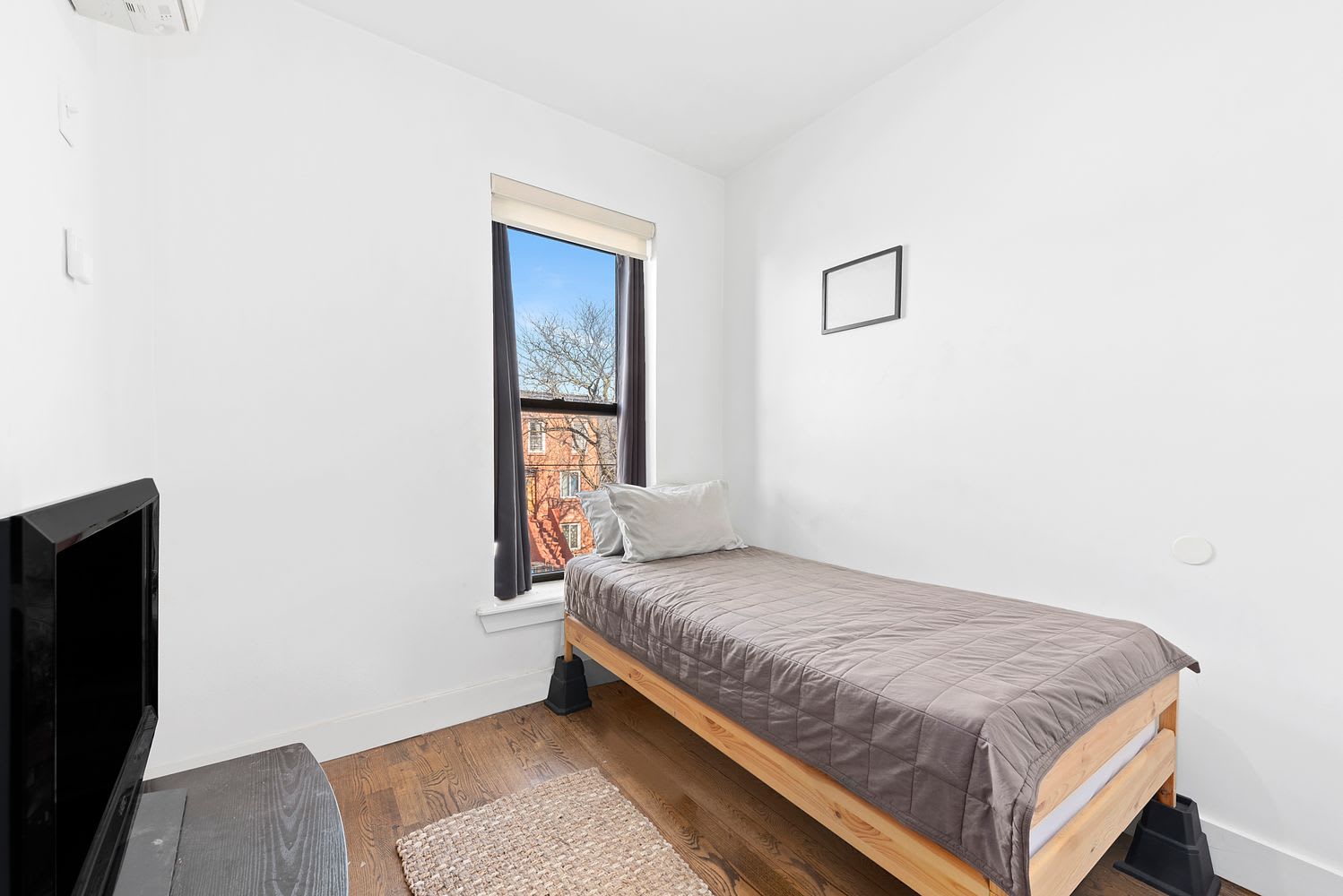 Bedroom with a wooden single bed, gray quilt, hardwood floors, and a tall window.