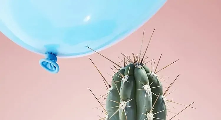 A blue balloon hovering above a cactus with a pink background. The image suggests a playful, precarious situation, likely metaphorical.