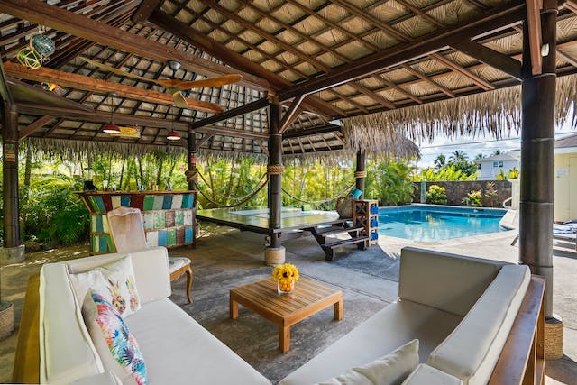 Covered outdoor living space with a gray sofa and a large swimming pool.