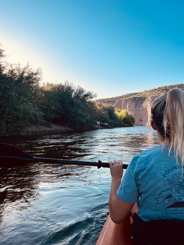 A person kayaking on a calm river, surrounded by lush greenery and cliffs under a clear blue sky.