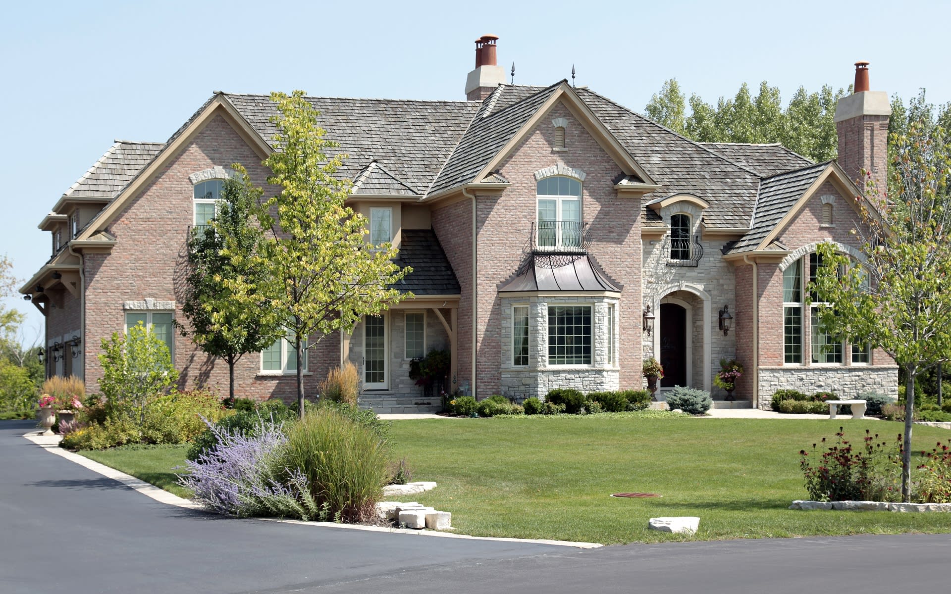 Large brick and stone house with gabled roofs, twin chimneys, and landscaped front lawn.