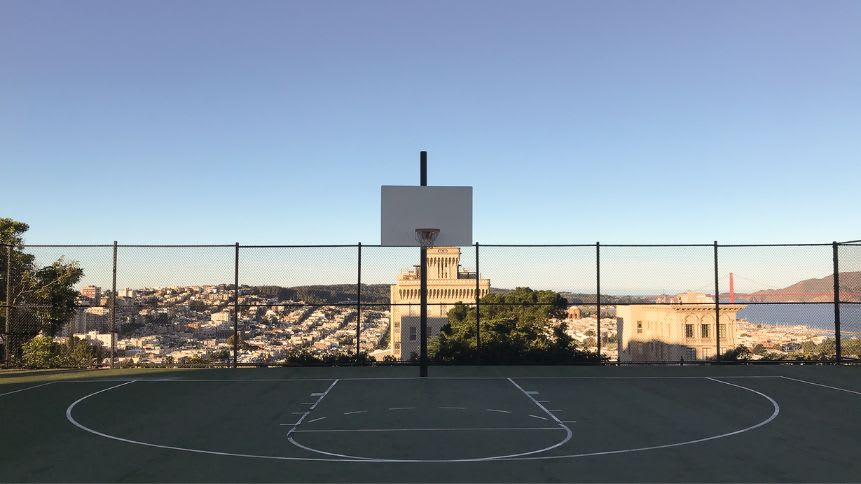 An outdoor basketball court at Alice Marble Tennis Courts in Russian Hill. The court is enclosed by a high chain-link fence. Beyond the court, there’s a stunning view of San Francisco’s cityscape, bathed in sunlight, with the Golden Gate Bridge visible in the distance under a clear sky.